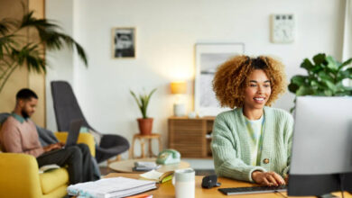 Smiling female freelancer using computer while colleague in background. Businesswoman is working at desk in home office. They are in apartment.
